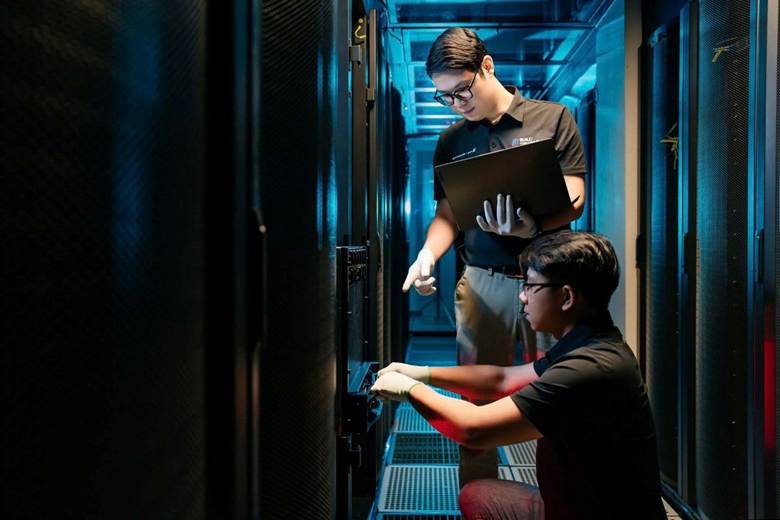 The image depicts two individuals collaboratively working on a computer server, which is housed within a server room with rows of rack-mounted equipment.
AI-generated content may be incorrect.